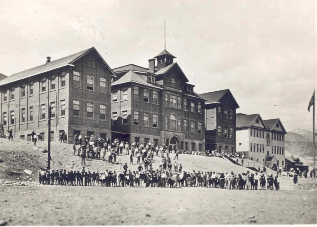 A historical black and white photo of Central Elementary School. The school building, captured likely in the early to mid-20th century, is a large, multi-story structure with prominent architectural features, including a central section with a peaked roof and a cupola. There are two wings flanking the central structure, each with rows of evenly spaced windows, indicative of classrooms. A group of students and perhaps some staff members are gathered in front of the school, many standing in lines, suggesting the photo might have been taken during a school event or class photo day. The terrain in the foreground is barren and sloped, leading up to the school, emphasizing its prominent position. The background is open sky, with the faint outline of mountains or hills, which along with the building style, suggests a Western or rural setting. This image serves as a visual record of the educational infrastructure and community during that period.