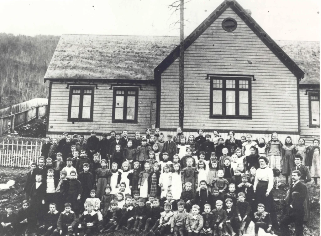 A historical black and white photograph of a school located on Green Avenue, situated on a bench above the townsite in Trail, British Columbia. The school, likely taken in the early 20th century, features a group of students and teachers posing for the photo outside the school building. The children are arrayed in several rows, with some sitting on the ground, others kneeling, and the rest standing. The teachers stand at the right end of the group. The students are dressed in various styles of the time, with many of the boys in knickers and shirts and the girls in dresses. The school building in the background is a simple wooden structure with visible windows, a shingled roof, and a bell tower, indicating its function as a place of learning. The surrounding area appears to be rural, with a fence running alongside and trees dotting the landscape behind. The image captures a sense of community and the educational setting of that era.