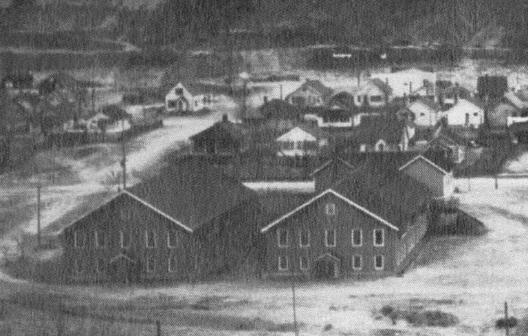 An old, grainy black and white photograph that captures East Trail School, with its distinctive gabled roofs, situated in a community setting surrounded by residential homes and a backdrop that suggests a small-town atmosphere. The image provides a glimpse into the school's role within the fabric of the neighborhood.