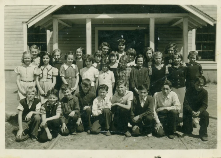 A black and white photograph from around 1932 of a group of students at Annable School. There are three rows of children, with girls mainly in the back and middle rows, and boys in the front, all positioned in front of the school's entrance. The students are dressed in the simple, practical attire of the era, with the boys in button-up shirts and slacks, some with ties, and the girls in a mix of dresses and blouses with skirts. A few students are kneeling in the front row, while the others stand straight and tall. Two female teachers stand amidst the students, wearing period-appropriate clothing. The children exhibit a range of expressions from smiles to serious gazes, providing a candid glimpse into the school life of that time. The school building behind them is modest, with a visible entrance and large windows, characteristic of early 20th-century rural school architecture.