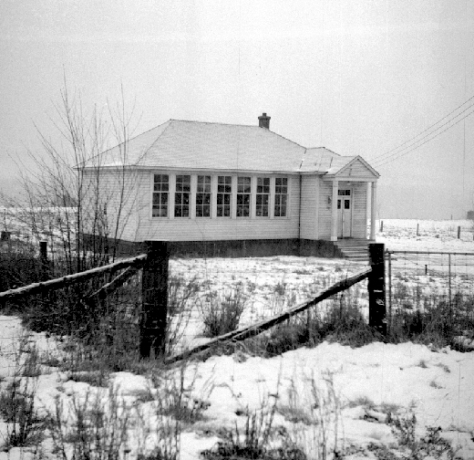 A black and white photograph of Beaver Falls School, with a view capturing its simple architecture amidst a snowy landscape, fenced by a rustic wooden barrier. The photo, attributed to the BC Archives, reflects a piece of educational history in British Columbia.