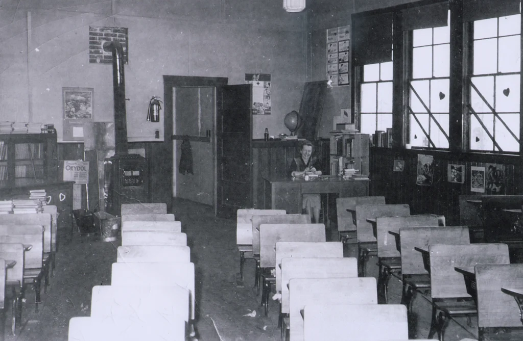 A black and white photograph of the interior of Casino School, featuring an early 20th-century classroom setting. Wooden desks arranged in neat rows face the teacher's desk at the front, where a young student sits, adding a personal touch to the scene. The room is heated by a prominent wood-burning stove near the center, a common fixture of the time. Large windows with diamond-shaped panes provide ample natural light, reflecting off the wooden floors. Educational posters and a world globe suggest a focus on geography and history, while the room's high ceiling and dark wood cabinetry exude a sense of the era's educational environment. The simplicity and order of the classroom highlight the utilitarian approach to education of that period.