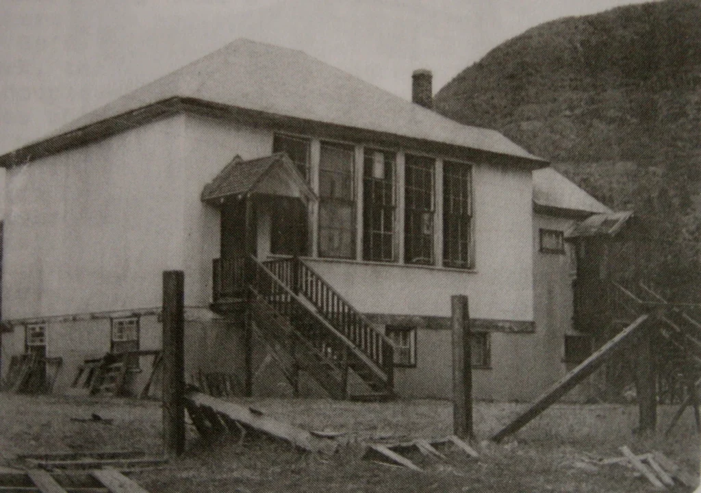 A black and white photograph of Columbia Gardens School, featuring its simple architecture with a peaked roof and rows of windows. A set of wooden stairs leads up to the main entrance, which is sheltered by a small porch. The building sits on a rough landscape with unfinished wooden beams and debris scattered in the foreground, suggesting either construction or disrepair. The stark, functional design is characteristic of rural schools from the early 20th century. Behind the school, the gentle slope of a hill rises, hinting at the school’s setting in a valley or rural area.