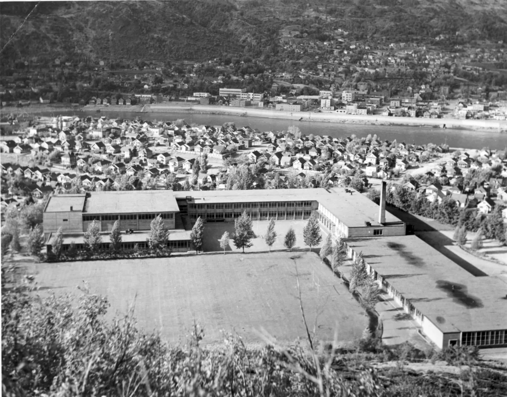 A black and white photograph of Crowe High School, taken from an elevated perspective showing the sprawling school complex with its distinctive mid-century architecture, large windows, and flat roofs, overlooking a densely populated residential area and the river in Trail, BC, encapsulating the school's prominence within the community.