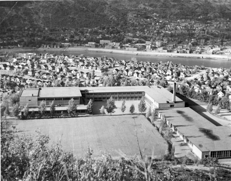 A black and white photograph of Crowe High School, taken from an elevated perspective showing the sprawling school complex with its distinctive mid-century architecture, large windows, and flat roofs, overlooking a densely populated residential area and the river in Trail, BC, encapsulating the school's prominence within the community.