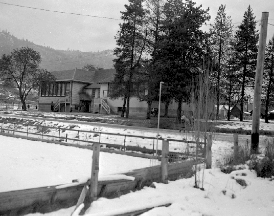 A black and white archival photograph of Fruitvale Elementary, showing a two-story, traditional wooden schoolhouse, with a backdrop of gentle hills and a foreground of snowy grounds, enclosed by a rustic wooden fence. This image is attributed to the BC Archives.