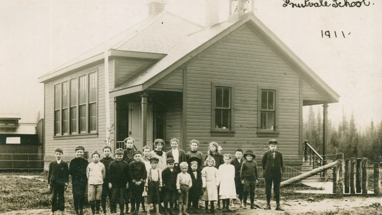 A sepia-toned historical photograph of the Fruitvale School from 1911. In front of the wooden schoolhouse stands a group of students, a mix of boys and girls of various ages, dressed in the style of the early 20th century. The children are lined up for the photo, with some seated on the ground. Behind them, the school features a prominent bell tower and large windows. The environment around the school is rural with trees and open land visible in the background, indicative of the school's community setting at the time.