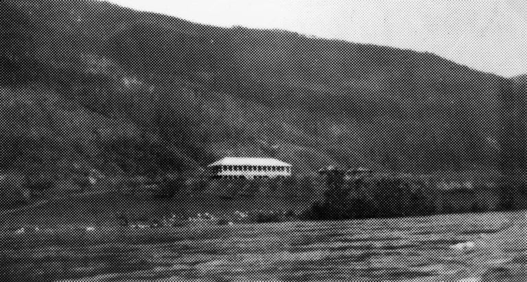 A grayscale image showing Glade School, a solitary building nestled on a hillside. The school's architecture is simple, with a gabled roof and visible windows. The surrounding landscape is mountainous and wooded, indicating a rural setting. In the foreground, the patterned texture suggests a body of water, adding a sense of isolation and tranquility to the school's location.