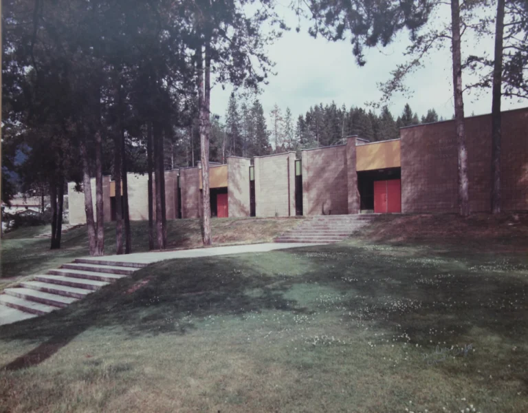 Photograph of the Beaver Valley Junior Secondary School taken during the day, showcasing its modern 1980s architecture. The building has clean lines, flat roofs, and a mix of brown and beige exterior walls, with a bold red door serving as the main entrance. A series of concrete steps lead up to the school, flanked by lush green grass and tall pine trees that imply a peaceful natural setting around the educational facility.