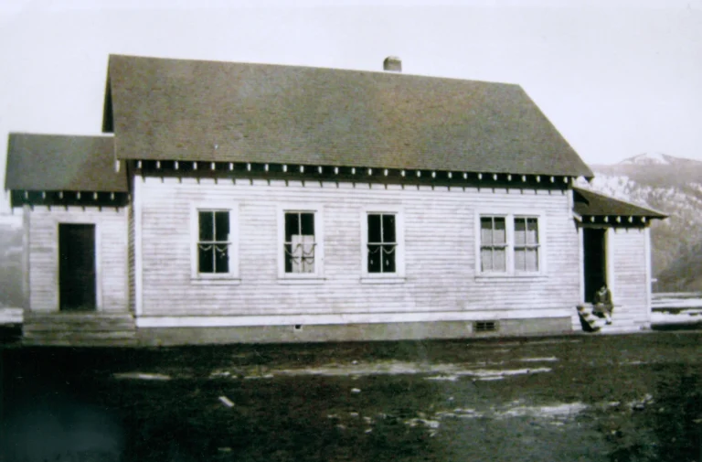 A historical black and white photo of the Kanamore School from 1927, showcasing its simple yet functional architecture with a gabled roof and multiple windows, typical of rural schoolhouses of that era, standing solitary in a clearing with mountains faintly visible in the background, symbolizing the roots of community education in a pastoral setting.