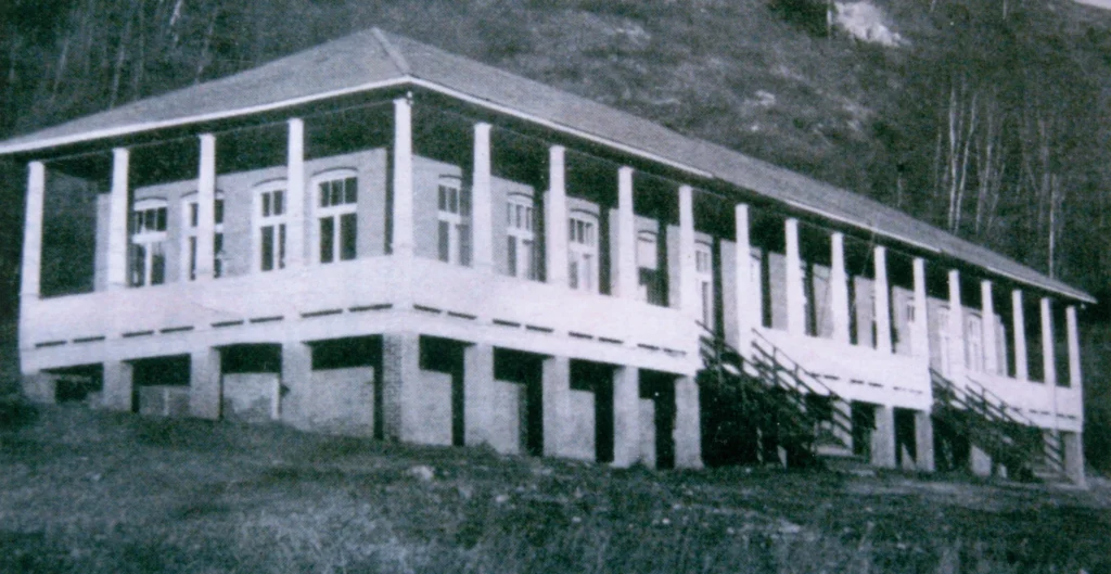 A grainy black and white photograph from 1934 of Raspberry School, portraying a two-story building raised on pillars with a series of steps leading up to the entrance, set against the backdrop of a forested hillside, typical of the rural school architecture of that time.