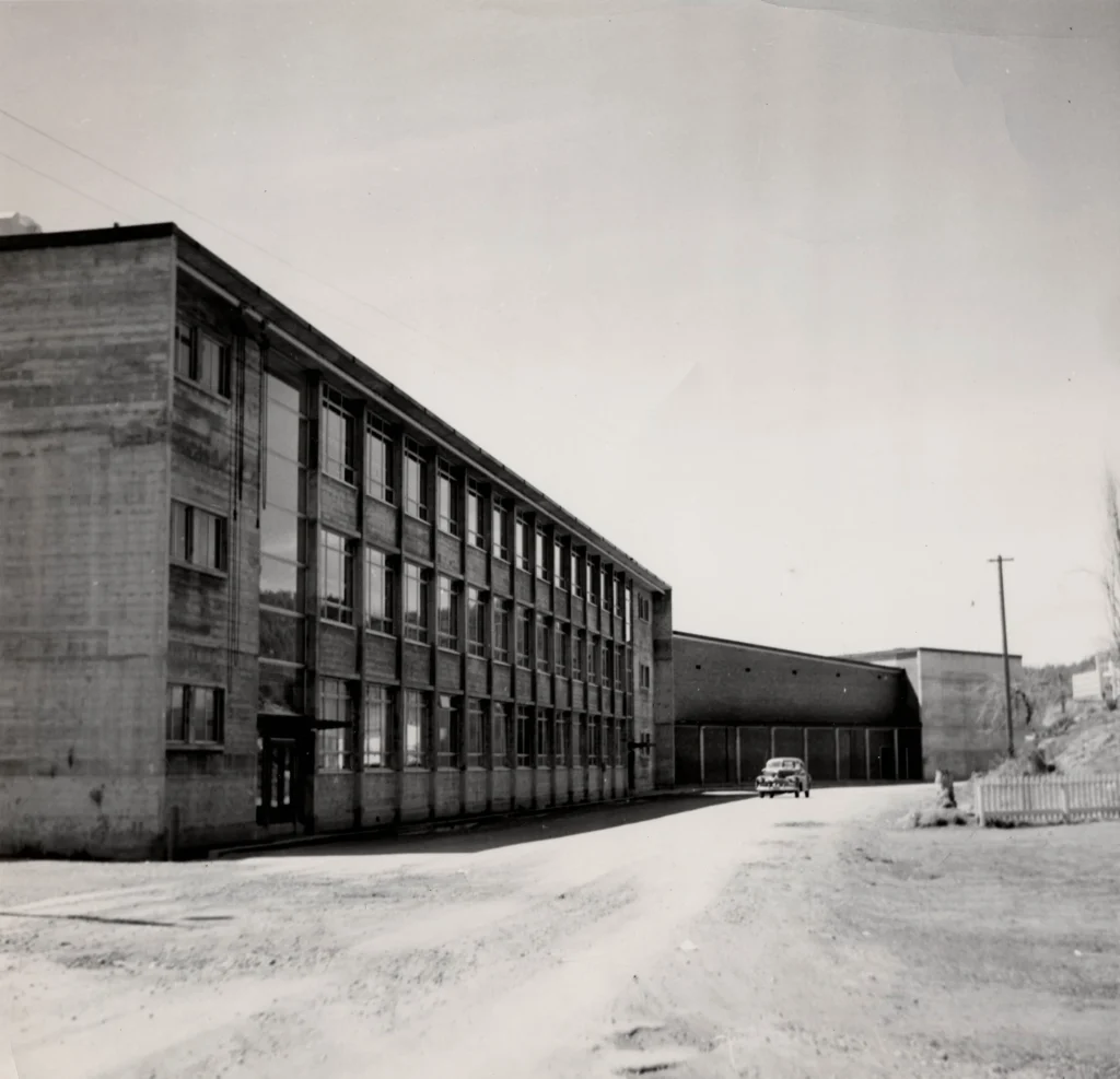 A black and white photograph from 1952 showcasing Rossland High School, a multi-story, rectangular building with long rows of windows, reflecting the utilitarian architectural style of the era, set on a street corner with a sparse landscape in the background.