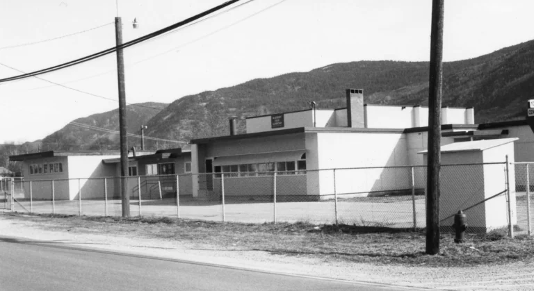 A black and white photo showing Blueberry Creek School, a single-story building of the late 20th-century design, secured behind a chain-link fence and set against the natural backdrop of a mountainous landscape, capturing the essence of a school located in a rural or suburban setting.