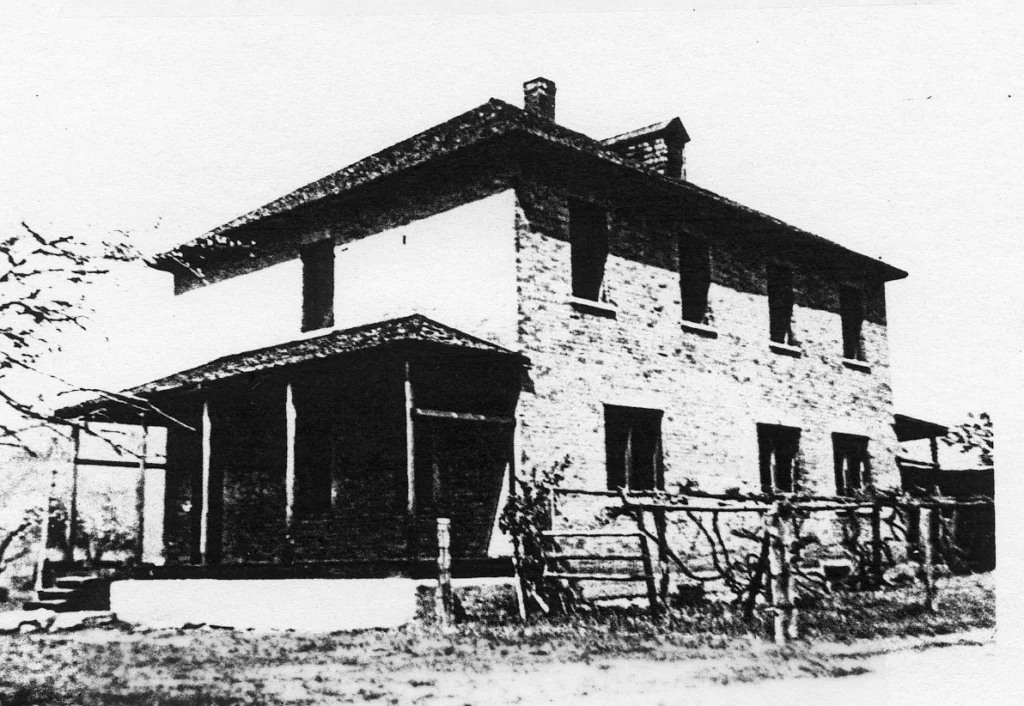 A stark black and white image of Brilliant No. 3 School, displaying a two-story brick building with a covered porch, characteristic of a robust, turn-of-the-century schoolhouse design, nestled within a pastoral setting.