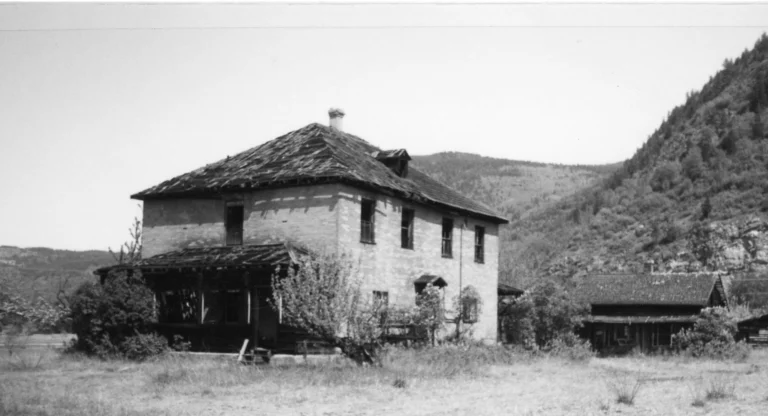 A black and white photo of Cay Creek School, an abandoned two-story brick building with a deteriorating roof and overgrown surroundings, standing as a relic of the past against the rugged hillside, illustrating the forgotten vestiges of early rural education.