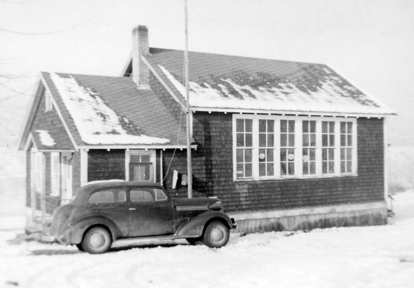 A nostalgic black and white photograph showing Champion Creek schoolhouse, with a classic vintage car parked out front. The snow on the ground and rooftop adds to the historical ambiance of the scene, harking back to an earlier time in educational history.