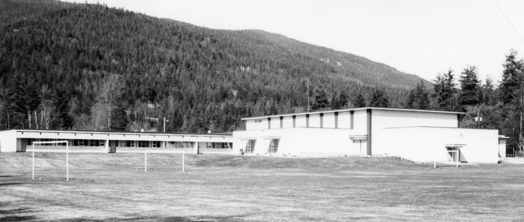 A black and white photograph from 1992 depicting Kinnaird Middle School, a modern building with a linear design, featuring a spacious sports field in the foreground and situated against a dense forest backdrop, exemplifying the integration of education and nature in school settings.