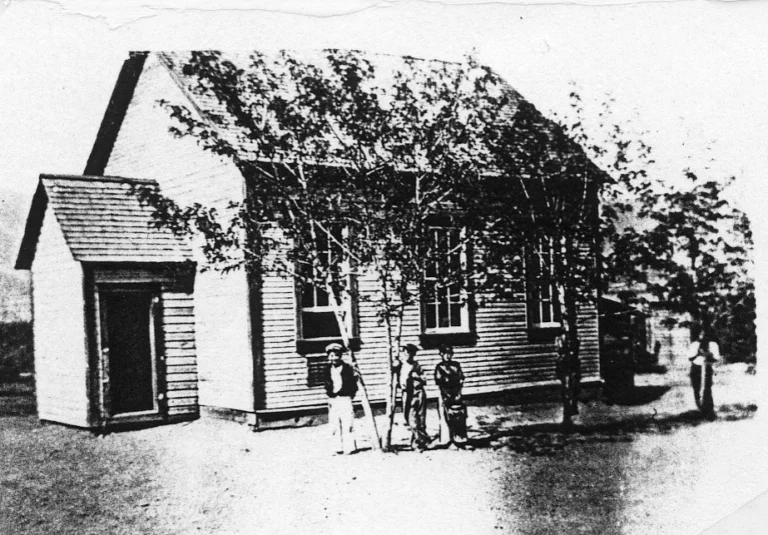 A grainy historical photograph of Kinnaird Stewardsville School, featuring a simple wooden structure shaded by trees, with children standing in front, conveying the rustic and communal atmosphere of early schooling in smaller communities.