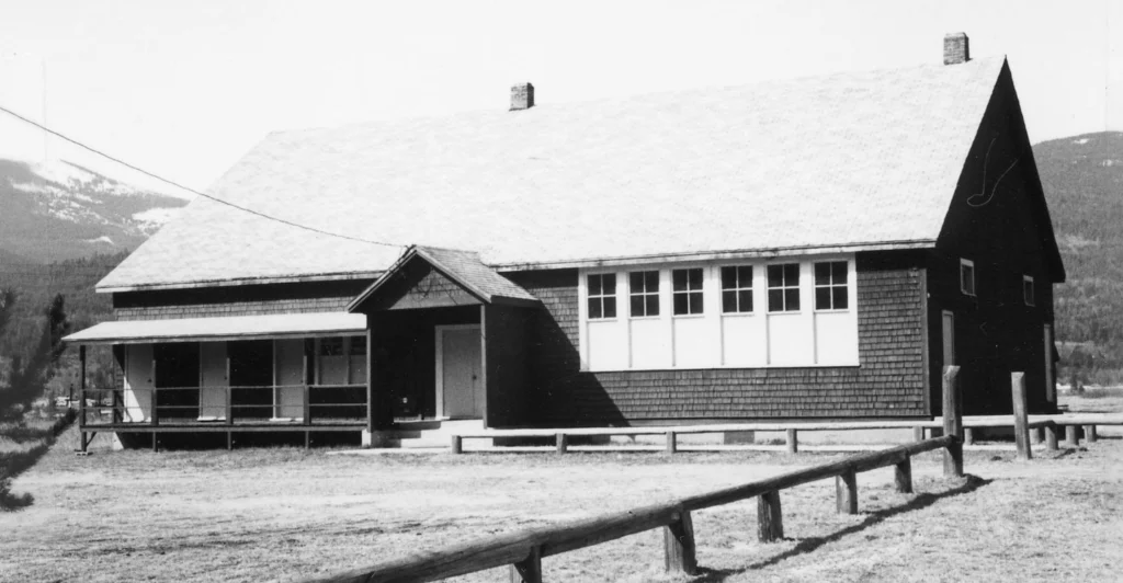 A black and white photograph of Ootischenia School, showing a quaint, gabled building with a covered porch, typical of early rural schoolhouses, set against a backdrop of mountainous terrain indicative of its location and the era of simple, community-based educational structures.