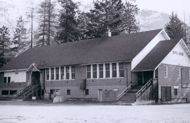 A vintage black and white photograph depicting the exterior of Castlegar Primary School, featuring its distinct pitched roof and the multiple entrance steps. The building is set against a backdrop of sparse trees and a lightly forested hill, evoking the rugged charm of historical school architecture in a natural landscape.