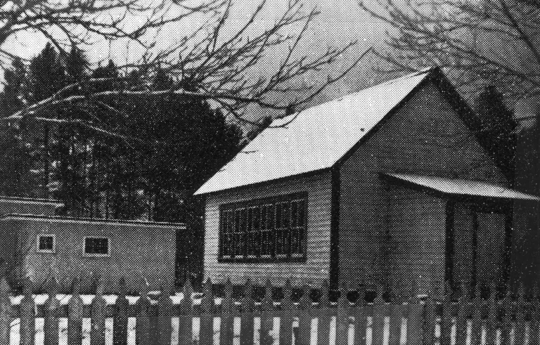 A halftone dot pattern effect black and white image of Renata School, showing a simple, small building with a pitched roof and a prominent window, fenced off by a white picket fence, indicative of small-scale, community-focused educational facilities.