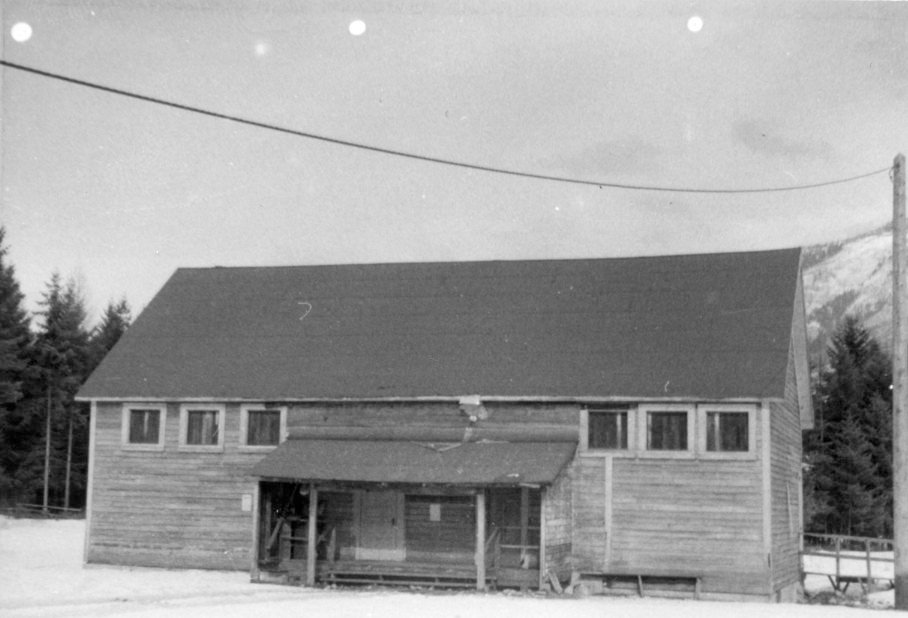 A black and white photograph of Thrums-Tarrys School, depicting a modest, two-story wooden building in a wintry setting, with snow on the ground and surrounded by tall trees, under a large expanse of sky with visible power lines, suggesting a quiet, rural educational setting from the past.