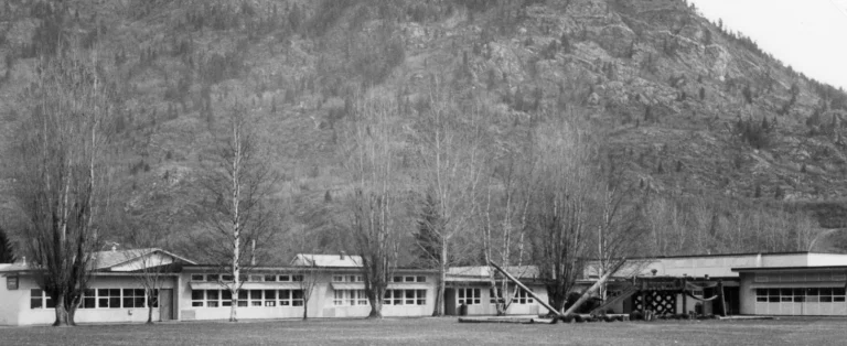 A black and white photograph from 1992 showing Twin Rivers School, a sprawling single-story educational building with large windows, situated in an expansive open field with a dramatic backdrop of forested mountains, indicative of the school's serene and natural setting.