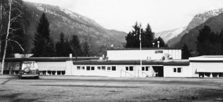 A black and white photograph of Woodland Park School, a low, extended building with a flat roof, situated in an open space with majestic mountains in the background, showcasing the natural beauty surrounding the educational facility.